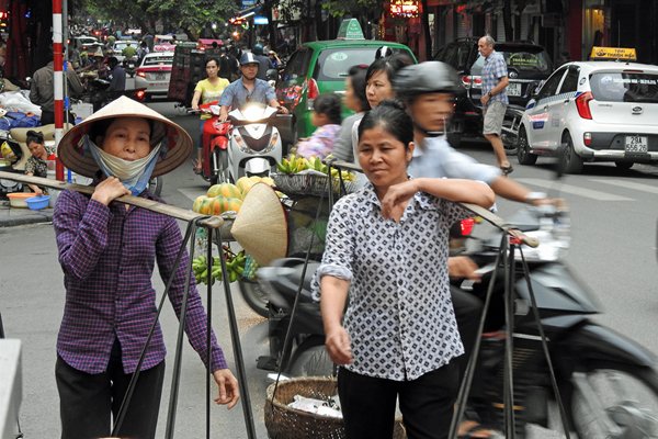 De een draagt zijn handeltje in Hanoi, de ander vervoert het op een brommer